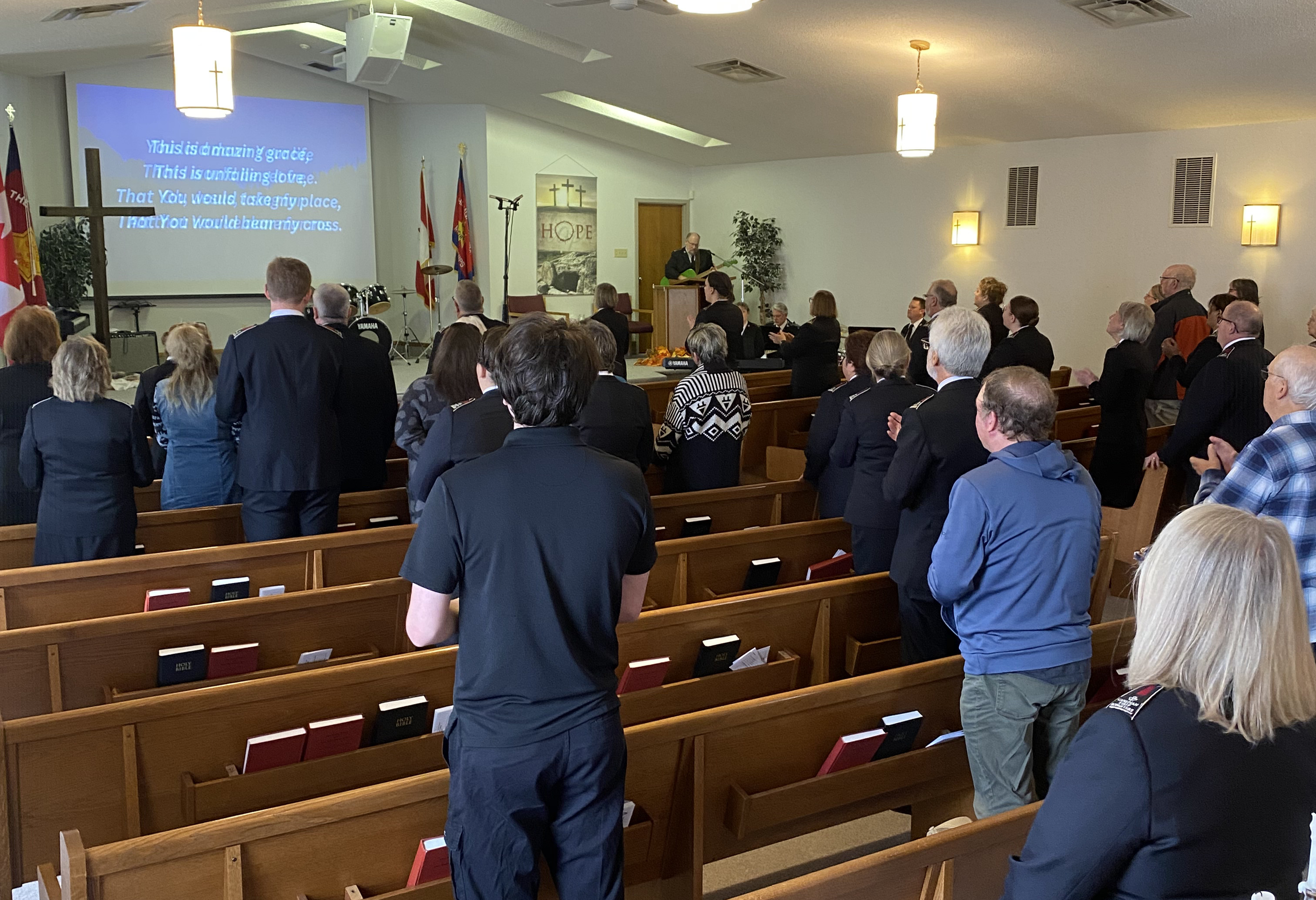 Congregation standing in wooden pews during a worship service, with lyrics projected on a screen at the front of the sanctuary.